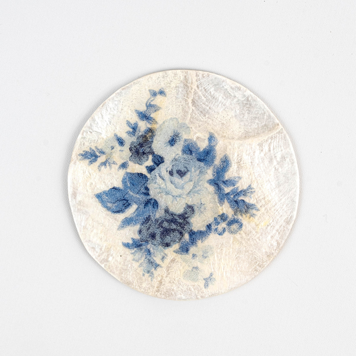 Handgjort underlägg i capizskal med blått blommigt decoupage, prytt med skira blå blommor mot en krämvit bakgrund — ett uttryck för hållbart hantverk och miljövänliga material