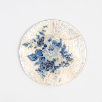 Handgjort underlägg i capizskal med blått blommigt decoupage, prytt med skira blå blommor mot en krämvit bakgrund — ett uttryck för hållbart hantverk och miljövänliga material