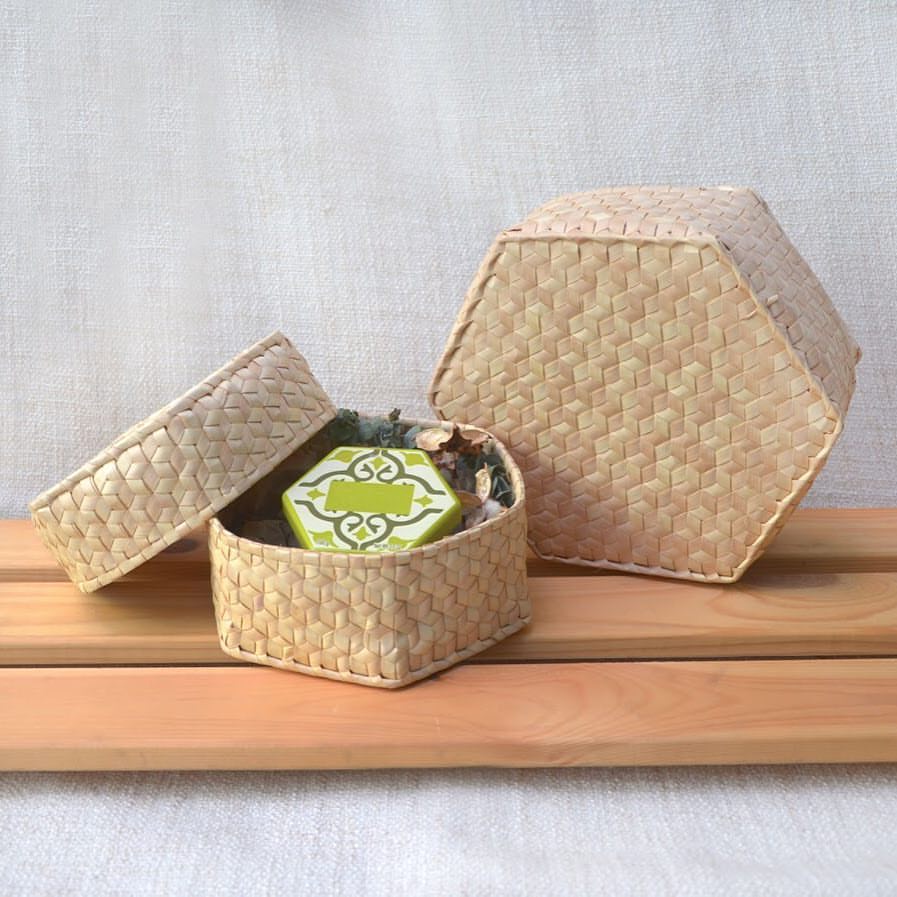 Käsintehdyt palmunlehtiset kuusikulmaiset rasiat luonnollisessa sävyssä, jotka esittelevät yksityiskohtaisia punontakuvioita. Yksi rasia on avoinna ja paljastaa sisuksensa, jossa on koriste-esine, korostaen Dese-tuotteen monipuolisuutta ja käsityön laatua.